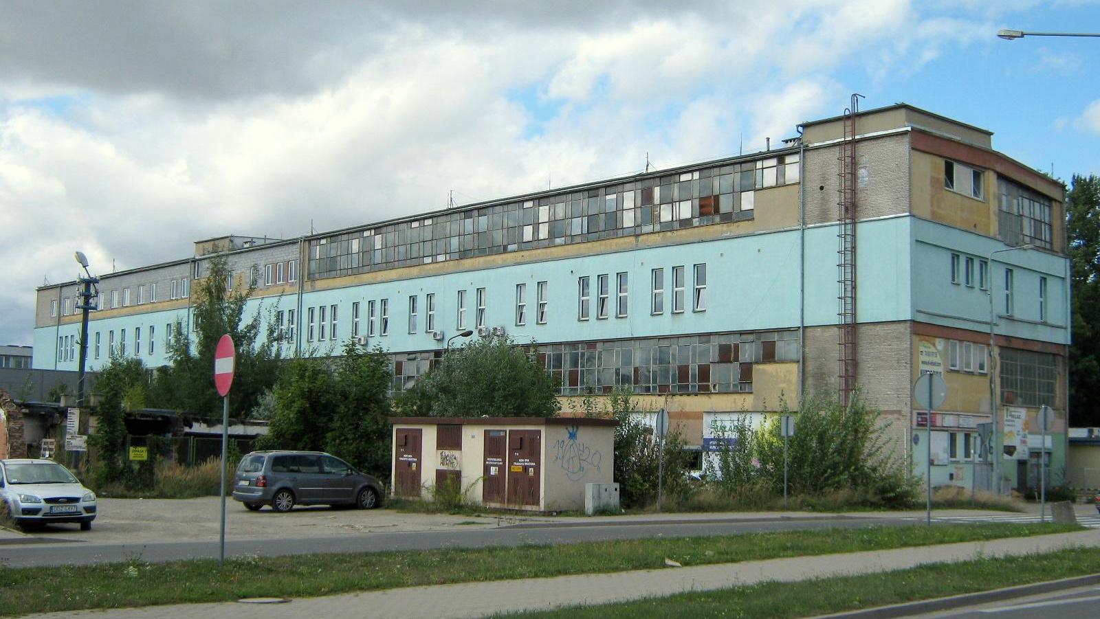 Trzypiętrowy budynek, z lewej strony parking z samochodami.