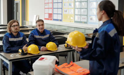 Kobieta w sali szkoleniowej trzyma żółty kask ochronny, naprzeciw niej siedzi dwóch mężczyzn w strojach roboczych. Na ławce leżą kaski, słuchawki i okulary ochronne oraz apteczka i kamizelka odblaskowa.