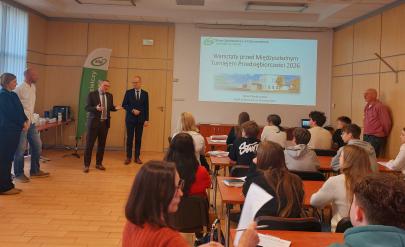 Rozpoczęcie warsztatów w sali szkoleniowej banku – po prawej stronie siedzą uczniowie i nauczyciele, po lewej organizatorzy, na wprost widoczny slajd prezentacji.