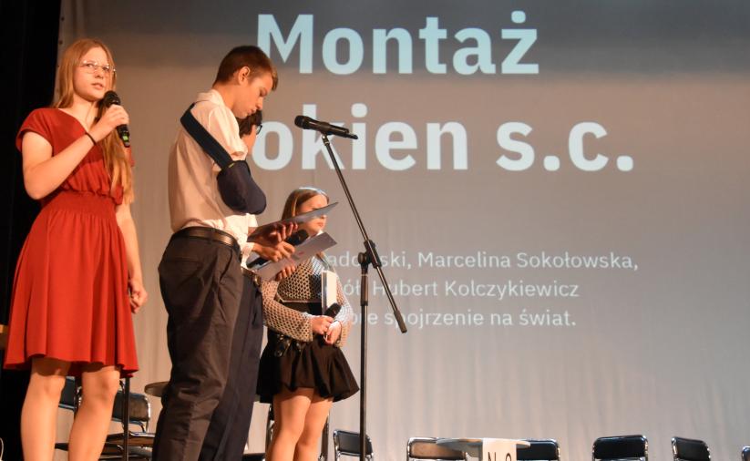 Uczniowie prezentują biznesplan na scenie, stojąc przy mikrofonach przed ekranem z tytułem prezentacji.