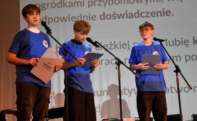Trzech uczniów stoi na scenie przed mikrofonami