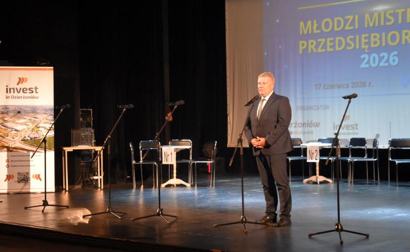 Burmistrz Dzierżoniowa przemawia na scenie podczas uroczystego otwarcia turnieju „Młodzi Mistrzowie Przedsiębiorczości 2026”. W tle widoczny jest ekran z nazwą wydarzenia oraz stanowiska dla uczestników.