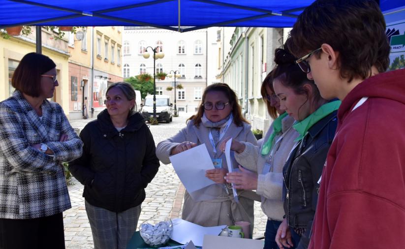 Grupa osób stoi pod niebieskim namiotem. W tle widoczny jest ratusz miejski w Dzierżoniowie.