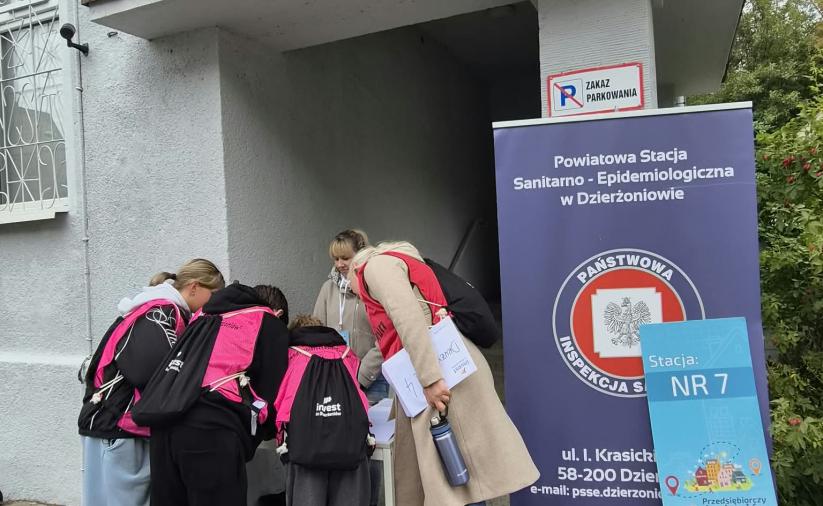 Czwórka uczestników gry w różowych kamizelkach widziana z tyłu, pochyla się nad stołem i rozwiązuje zadanie na stacji gry. Obok stoi opiekun drużyny oraz organizatorka stacji. W tle budynek.