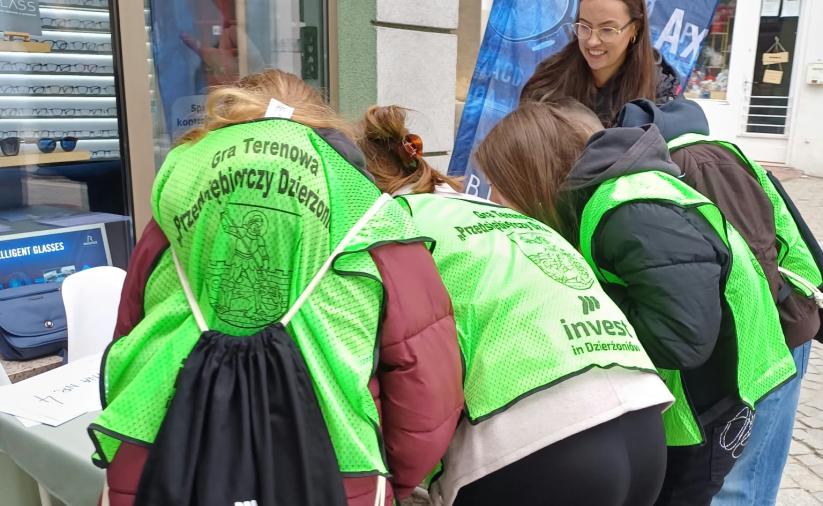 Czwórka uczestników gry w kamizelkach widziana z tyłu, pochyla się nad stołem i rozwiązuje zadanie na stacji gry. Obok stoi opiekun drużyny. 