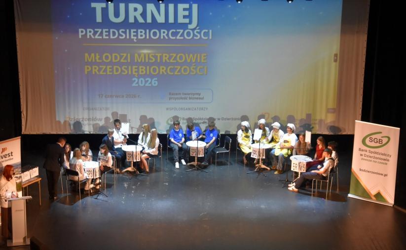 Dziecie siedzą przy 5 stolikach na scenie, w tle slajd Międzyszkolny Turniej Przedsiębiorczosci