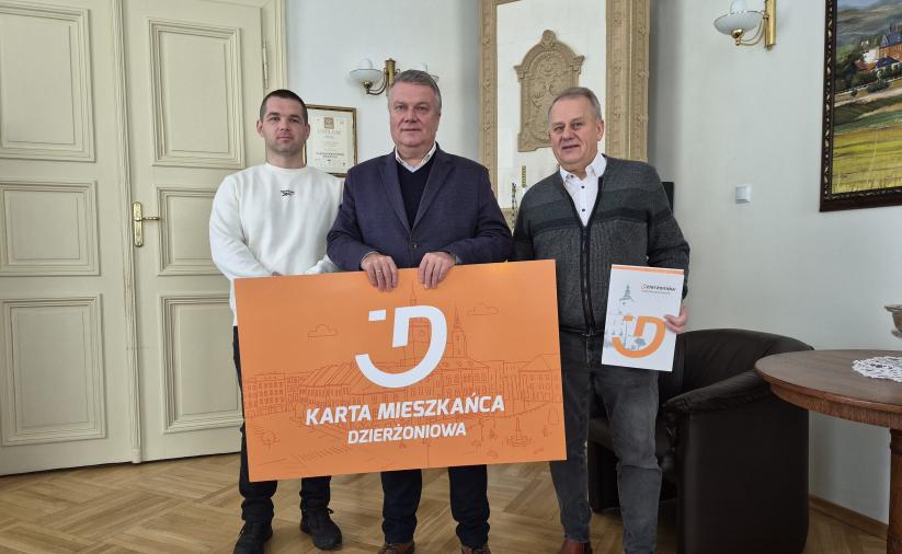 Burmistrz Dzierżoniowa i dwóch przedsiębiorców trzymają dużą pomarańczową planszę z napisem Karta Mieszkańca Dzierżoniowa.