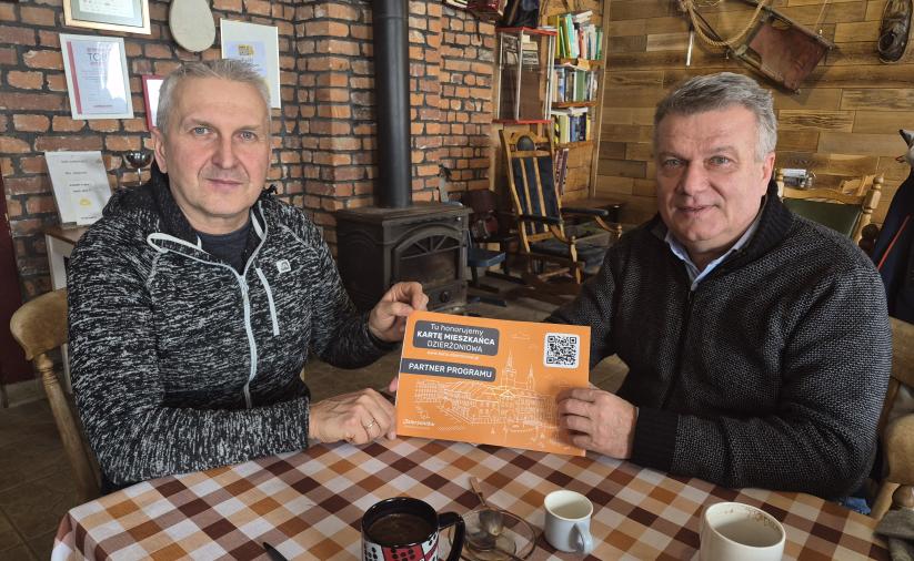 Dwóch mężczyzn przy stoliku trzyma naklejkę "Partner Programu. Tu honorujemy Kartę Mieszkańca Dzierżoniowa"