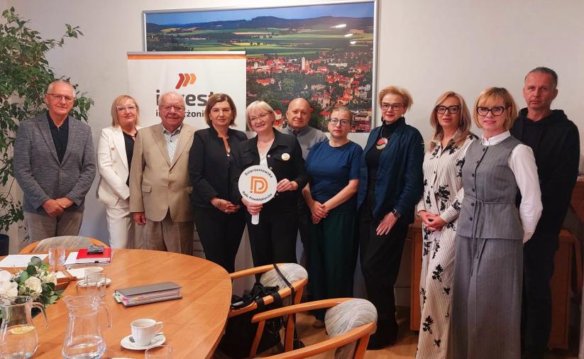 grupa członków Dzierżoniowskiej Rady Przedsiębiorców podczas spotkania w Urzędzie Miasta. Dziewięć osób stoi przy stole konferencyjnym, na tle panoramy Dzierżoniowa i baneru „Invest Dzierżoniów”. Pośrodku jedna z uczestniczek trzyma okrągły znak Rady Przedsiębiorców.