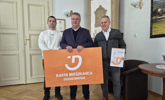 Burmistrz Dzierżoniowa i dwóch przedsiębiorców trzymają dużą pomarańczową planszę z napisem Karta Mieszkańca Dzierżoniowa.