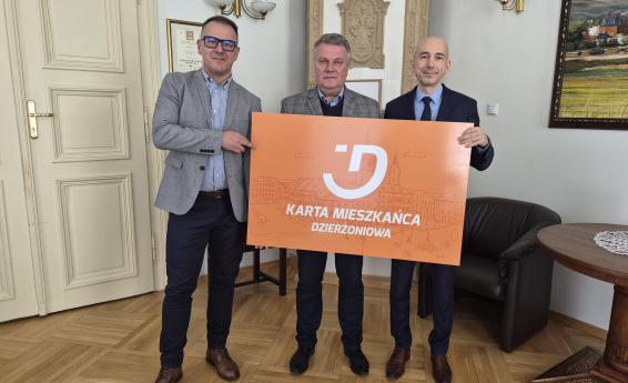 Three men standing in an office are holding a large orange Dzierżoniów Resident Card.