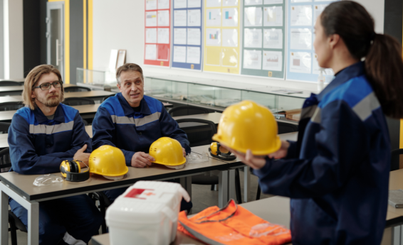Kobieta w sali szkoleniowej trzyma żółty kask ochronny, naprzeciw niej siedzi dwóch mężczyzn w strojach roboczych. Na ławce leżą kaski, słuchawki i okulary ochronne oraz apteczka i kamizelka odblaskowa.
