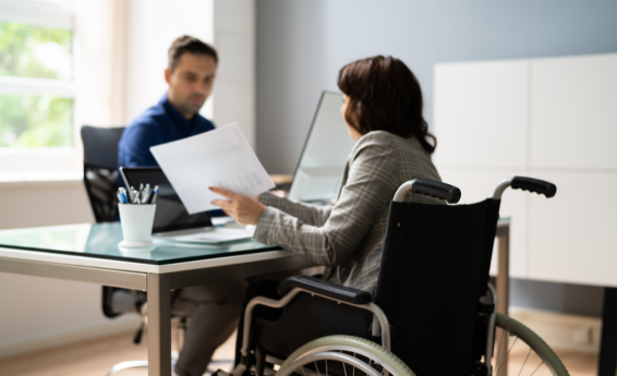 Kobieta poruszająca się na wózku inwalidzkim trzyma dokumenty, naprzeciw niej przy biurku siedzi mężczyzna.
