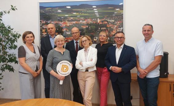 A group of seven people in a conference room, standing in front of a photograph of Dzierżoniów. One of the participants is holding a round logo of the Dzierżoniów Entrepreneurs Council.